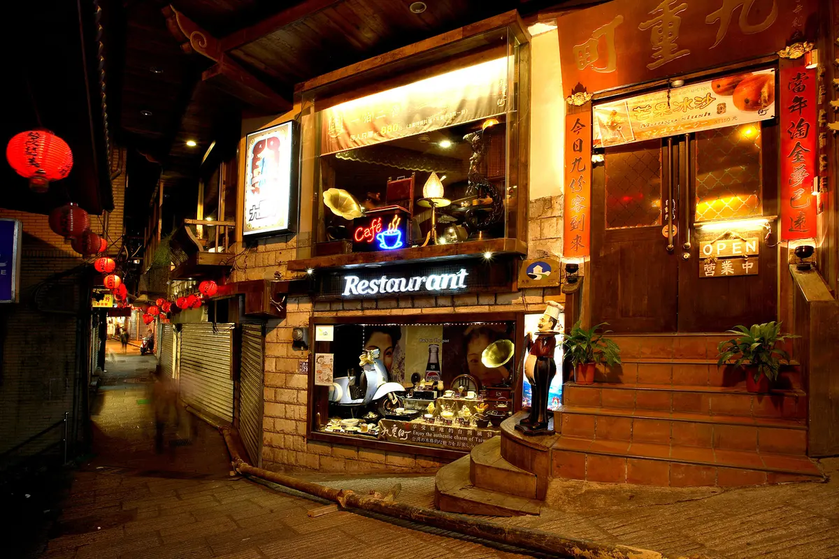 Un aparador en un carreró de Jiufen de nit, amb un aparador il·luminat, un rètol de cafè i fanalets de paper vermells penjats al llarg del carreró a l'esquerra