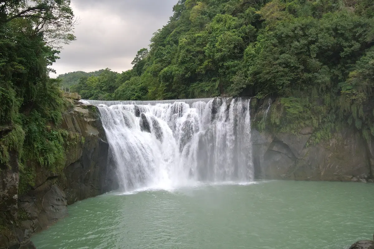 La cascada de Shifen, una àmplia cortina d'aigua blanca que cau sobre una cornisa rocosa cap a una gorga verda, amb vessants boscosos a banda i banda